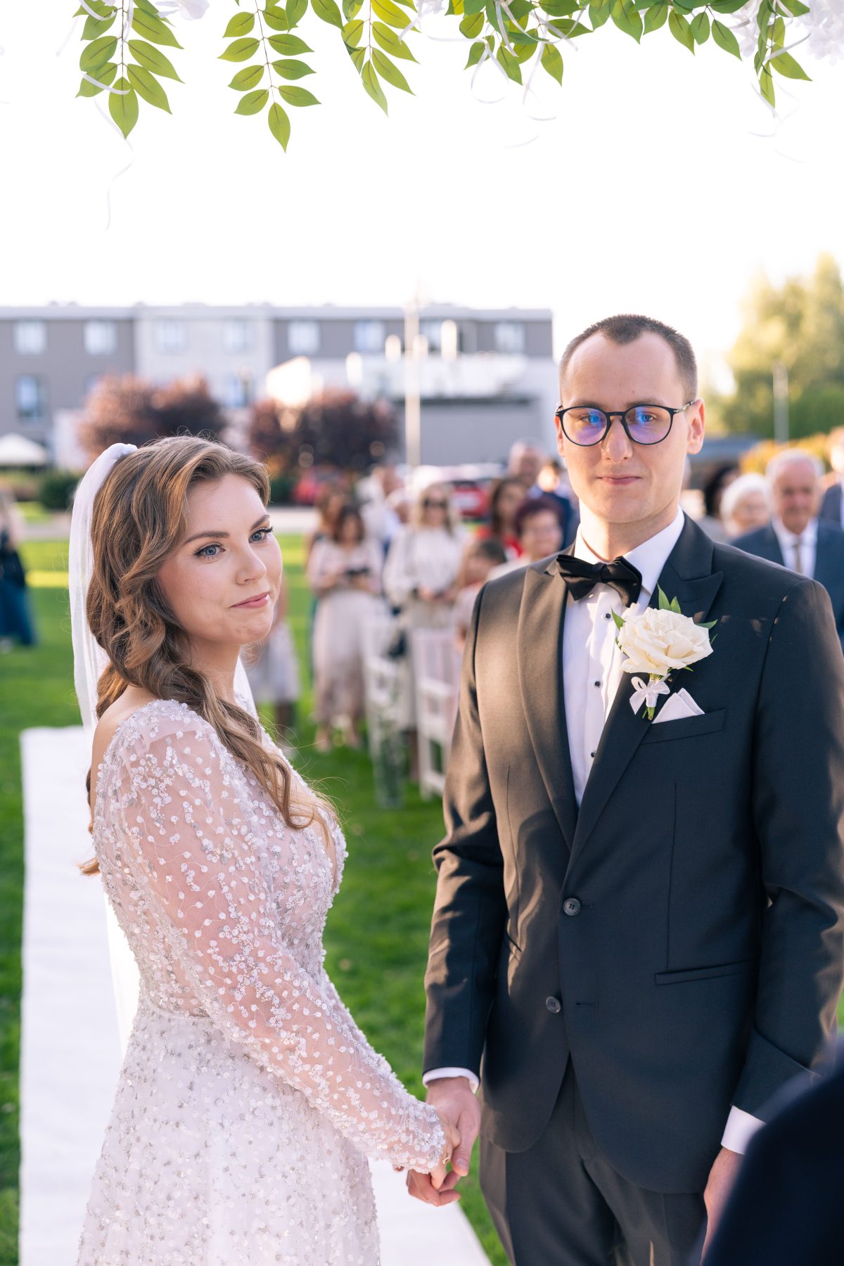 Para młoda trzyma się za ręce pod zielonym łukiem w trakcie ceremonii