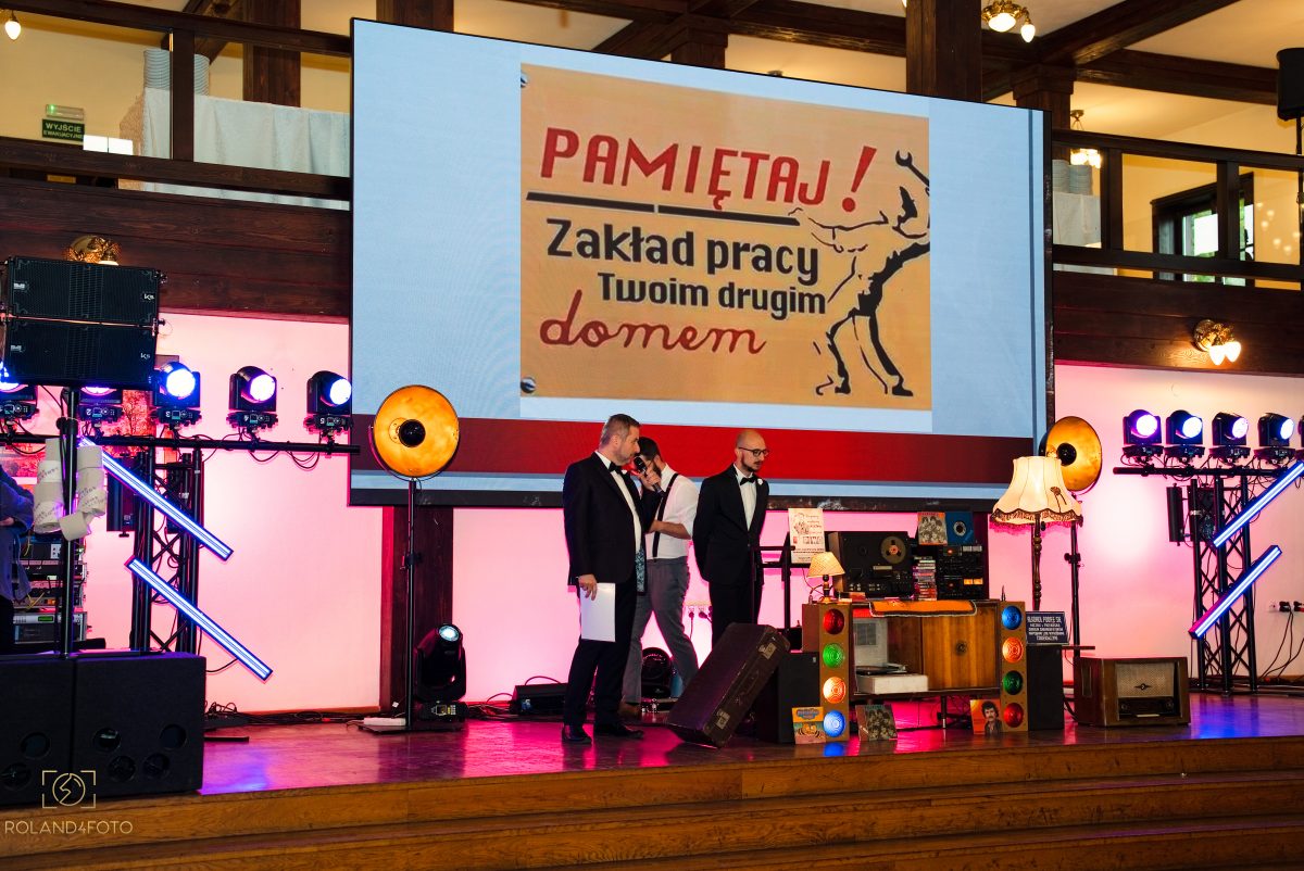 Ekran nad sceną z hasłem w stylu PRL: Pamiętaj! Zakład pracy Twoim drugim domem